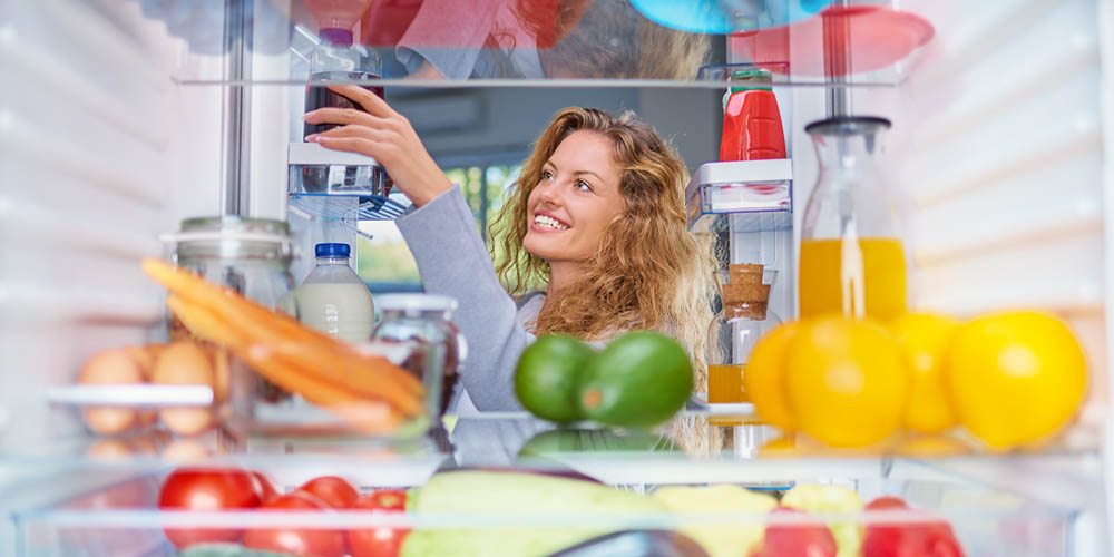 Blick durch einen gereinigten Kühlschrank, in dem verschiedene Gemüse auf Glasböden liegen. Im Hintergrund greift eine fröhliche lächelnde Frau nach einem Gegenstand in einem Türfach.